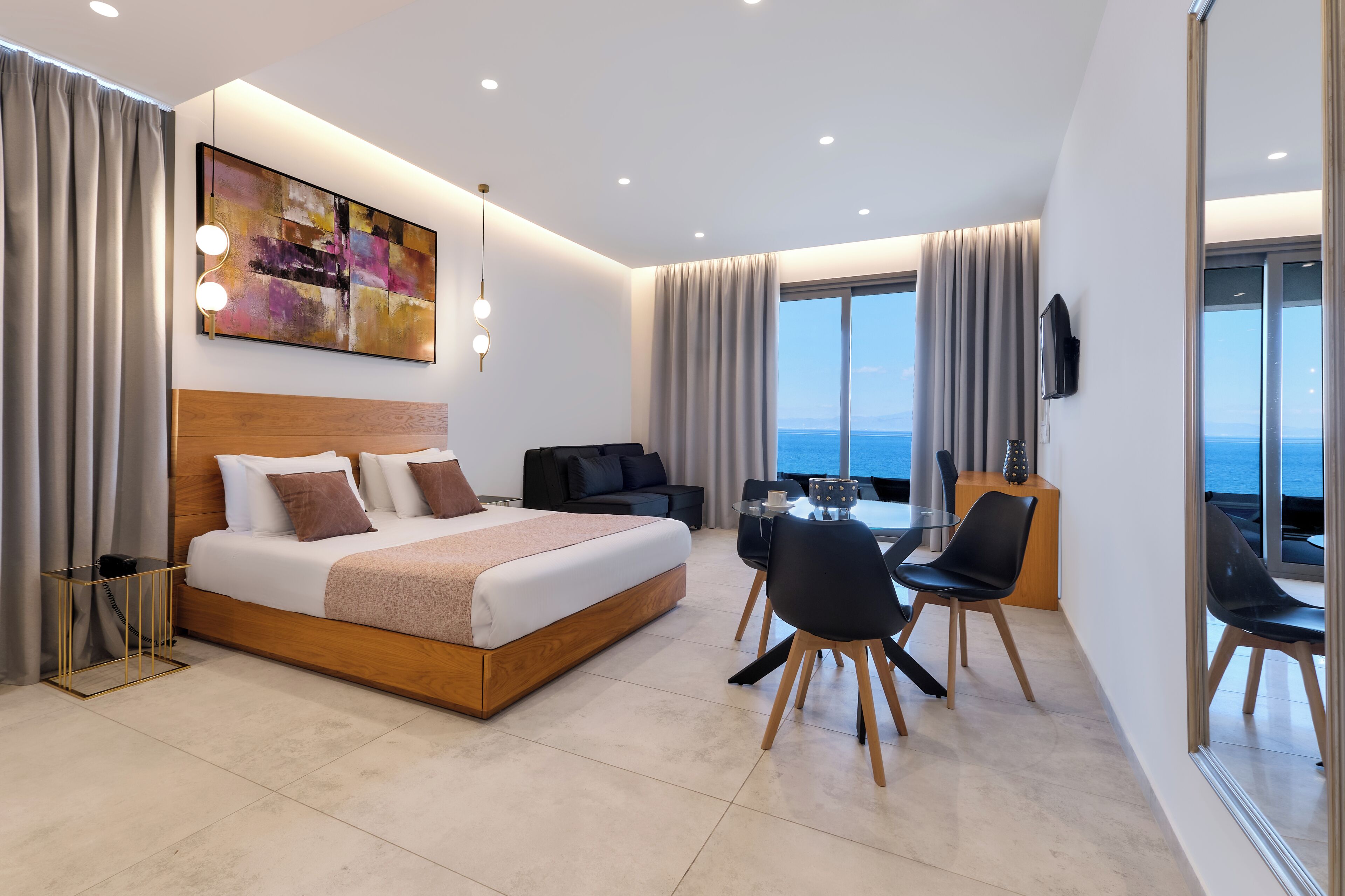 Habitación de hotel moderna con una cama grande, zona de comedor, televisión y vista al mar.