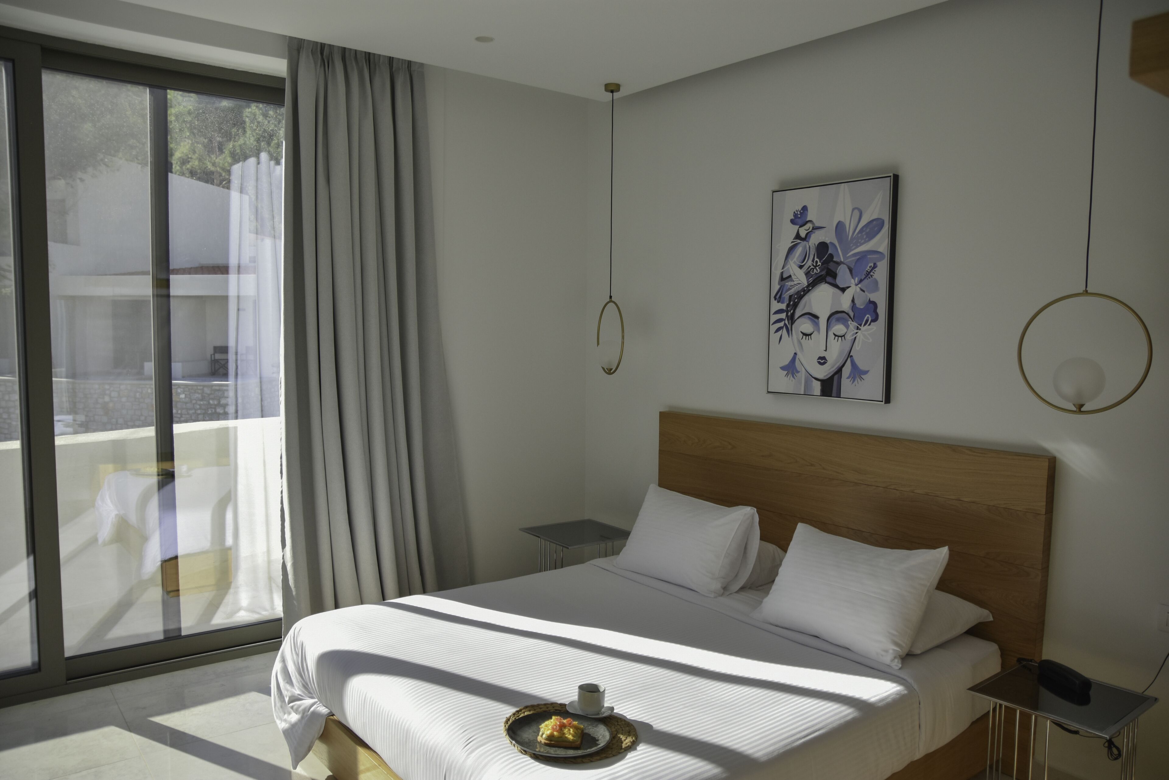 A modern hotel room with a large bed, a bedside table, a small table, and a view of the outdoors through a sliding glass door.