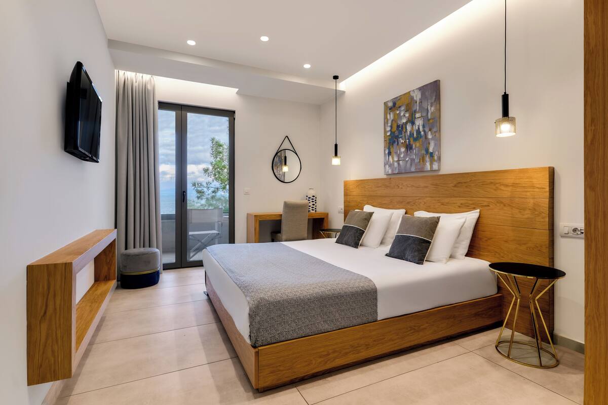 Ein modernes Schlafzimmer mit einem Holzkopfteil, einem großen Bett, einem Nachttisch, einem an der Wand montierten Fernseher und Blick ins Freie durch eine Schiebetür.
