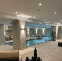 Un área de piscina interior moderna con una piscina central, múltiples chorros de agua y una zona de estar con sofás.