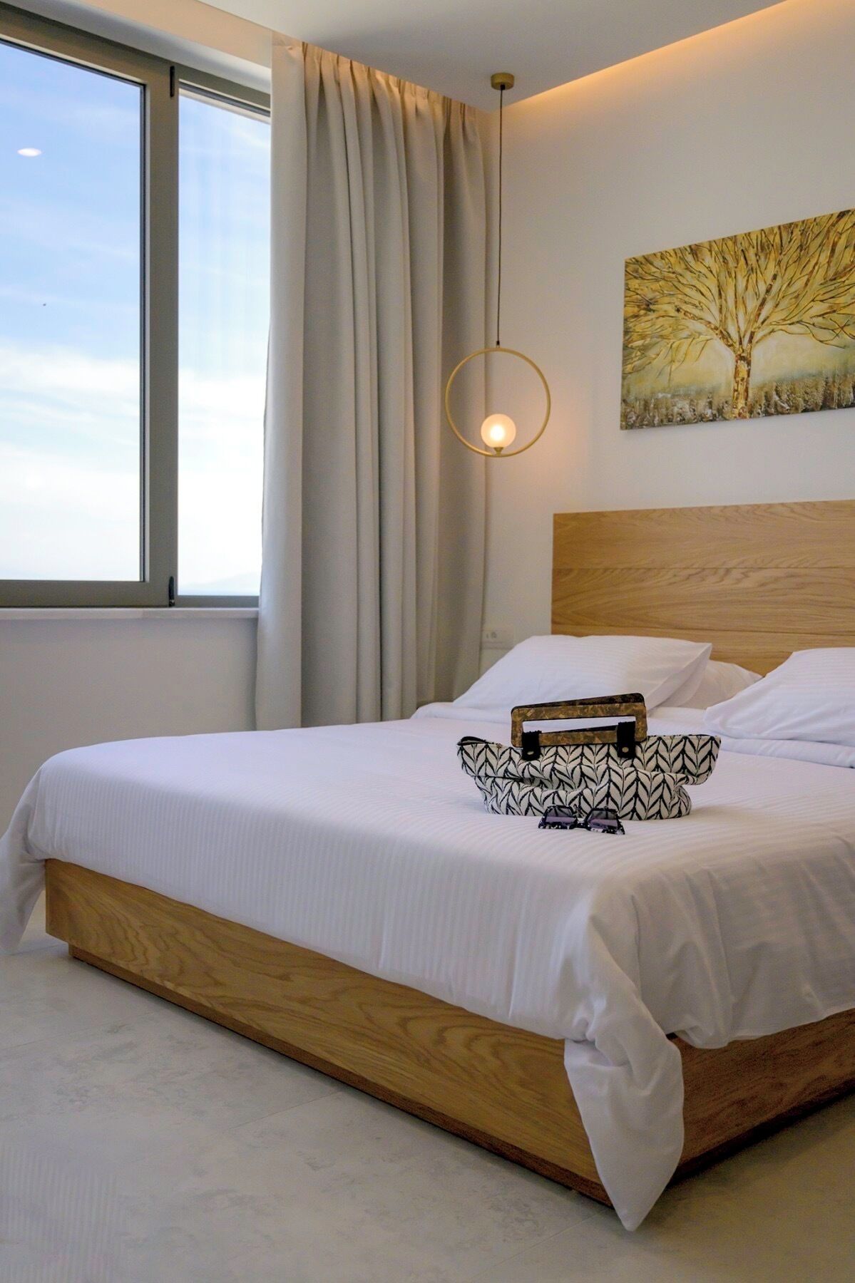 Una cama bien hecha con cabecera de madera, una mesita de noche con un bolso estampado, y un cuadro de un árbol en la pared.
