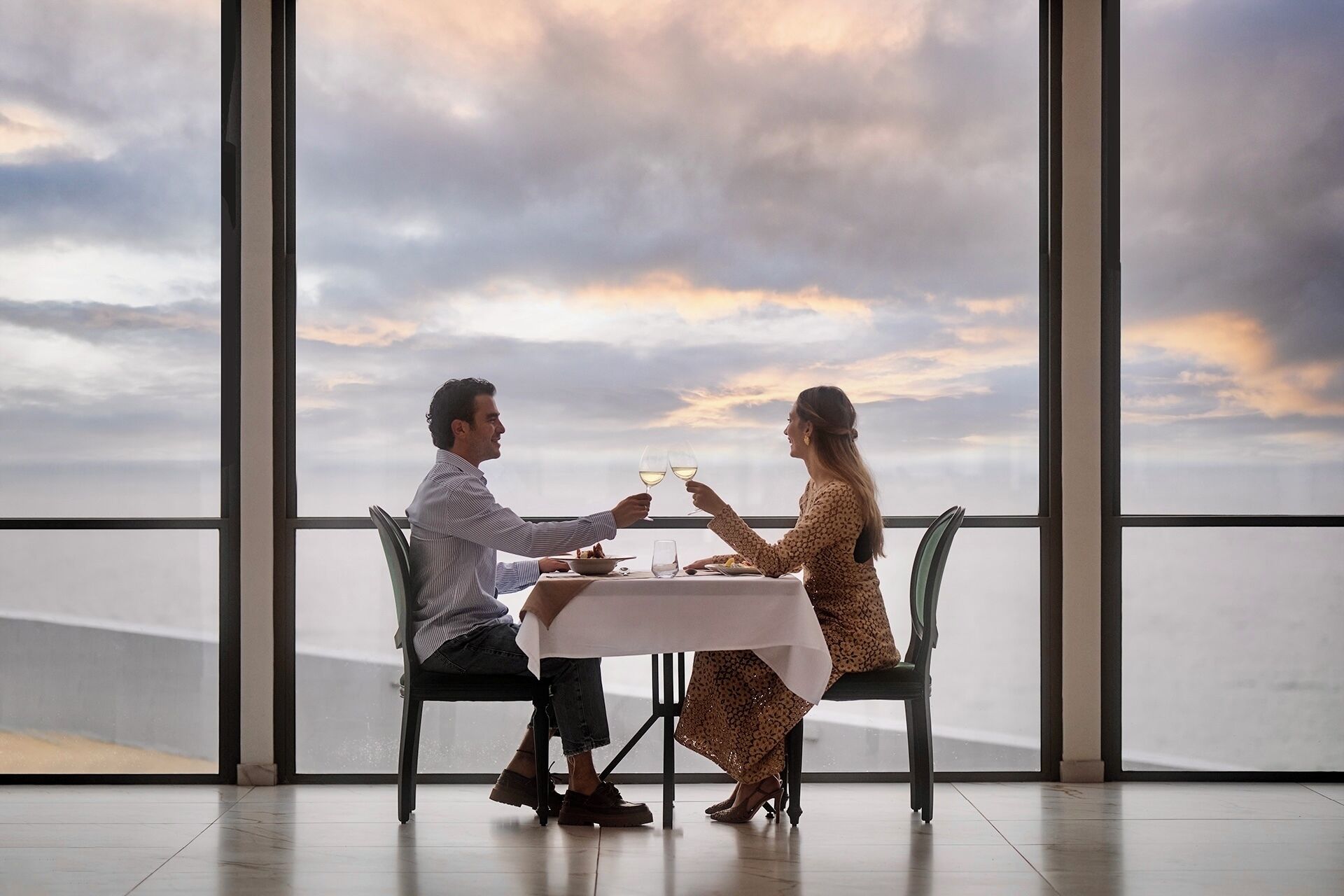 A couple dining at a table with a view of the ocean and sky through large windows.