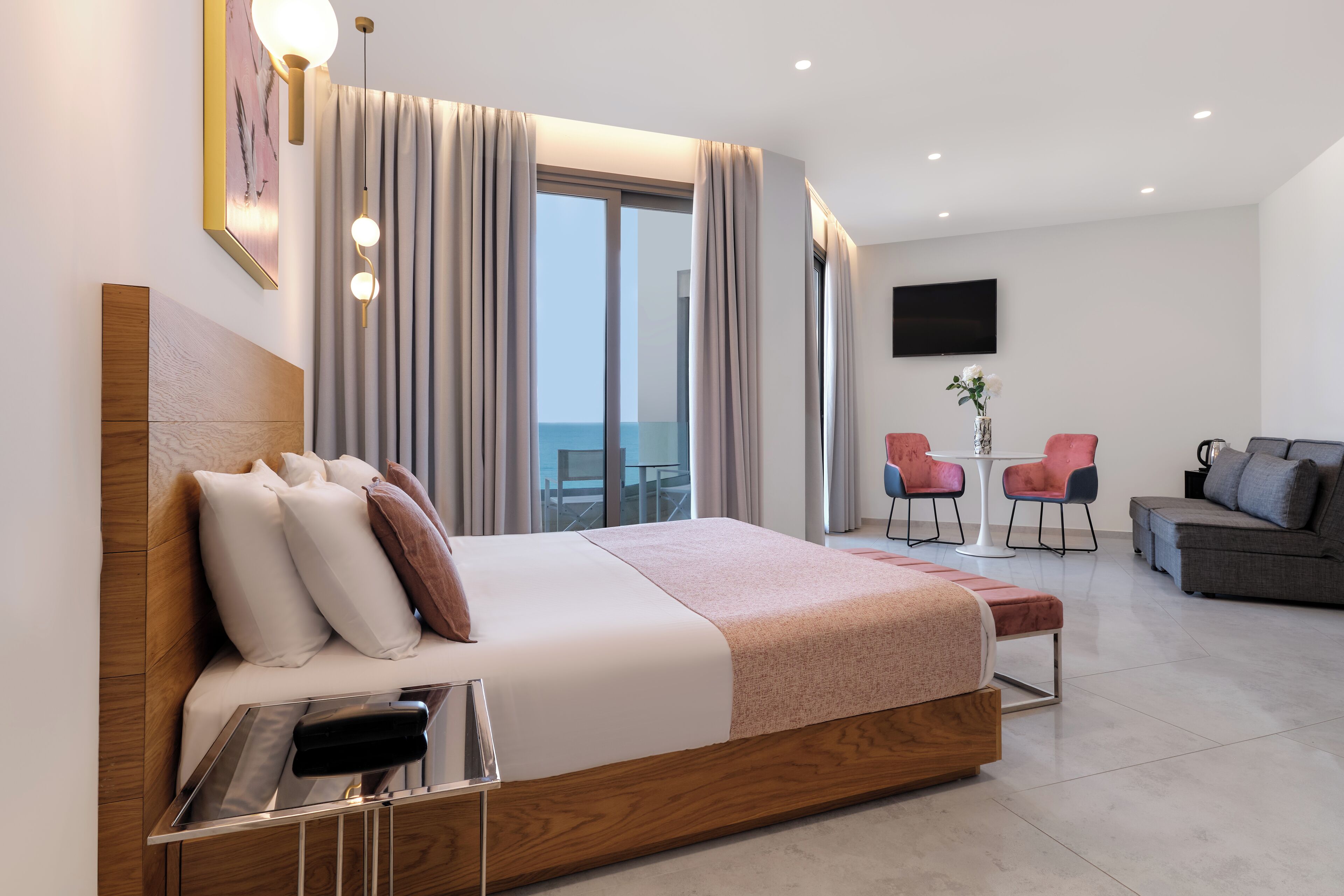A modern hotel room with a large bed, a flat-screen TV, a balcony with a view, and a seating area with chairs.