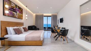 A modern hotel room with a large bed, a desk with chairs, a TV, and a view of the sea.