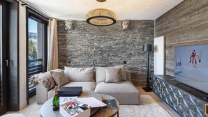 Un saló modern amb una paret de pedra, una gran televisió de pantalla plana, un sofà, una taula de centre amb una ampolla i una caixa, i una làmpada penjant.