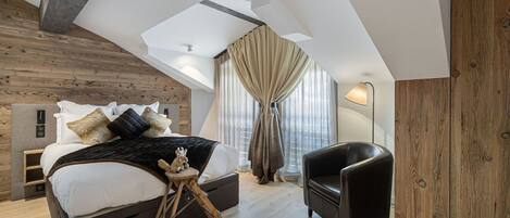 A modern bedroom with a loft-style ceiling, a bed with multiple pillows, a small wooden stool, a black leather chair, and a large window with curtains.