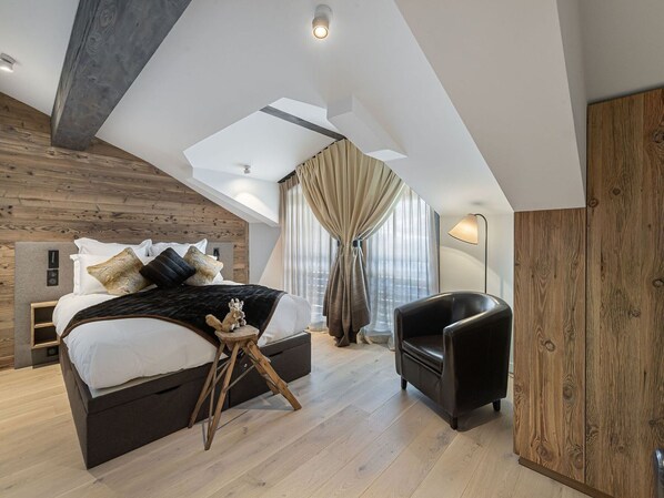 A modern bedroom with a loft-style ceiling, a bed with multiple pillows, a small wooden stool, a black leather chair, and a large window with curtains.