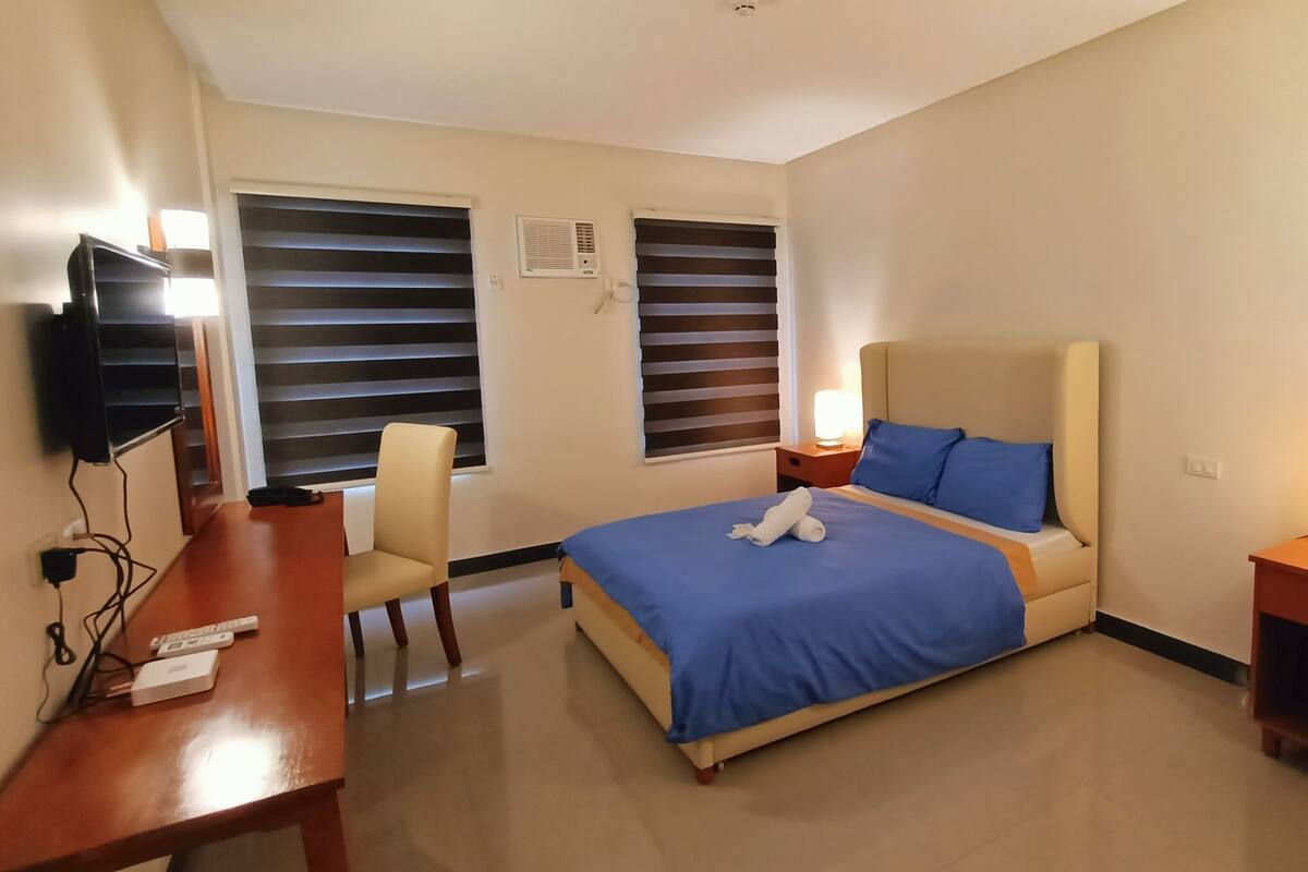 A hotel room with a bed, a desk, a chair, a TV, and two windows with blinds.