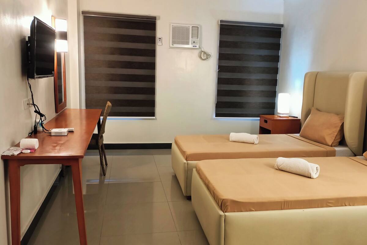 A hotel room with two beds, a desk, a chair, a TV, and a window with blinds.