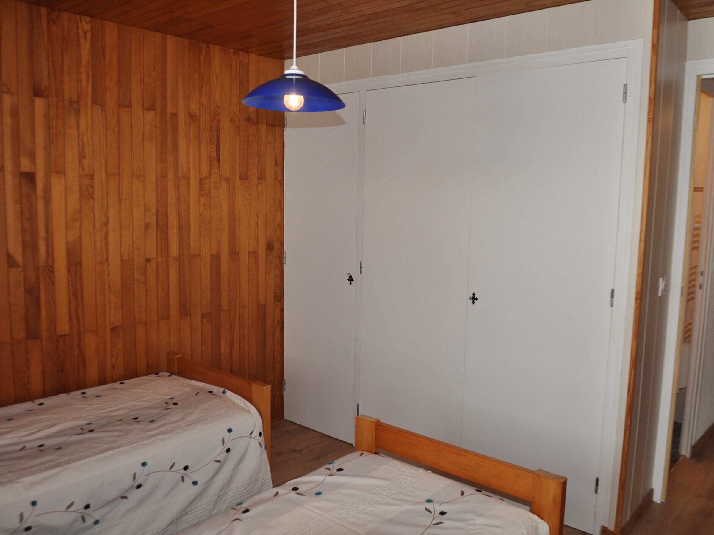 Une chambre avec deux lits simples, des murs recouverts de panneaux de bois, une armoire blanche et une lampe suspendue bleue.