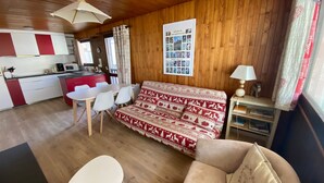 Une pièce chaleureuse avec un plafond en bois, un canapé recouvert d’un tissu à motifs, une table à manger avec des chaises, une petite table d’appoint avec une lampe, et un coin cuisine avec des placards rouges et un four à micro-ondes.