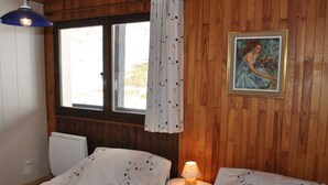 Ein Zimmer mit Holzwänden, zwei Betten mit blumigem Bettzeug, einem kleinen Tisch mit Lampe, einem Fenster mit Vorhängen und einem gerahmten Bild an der Wand.