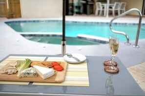 A poolside table with a cheese platter, a glass of champagne, and a plate with cutlery.