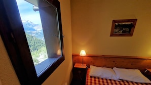 Une chambre avec un lit, une table de chevet, une lampe, une fenêtre donnant sur les montagnes et un tableau encadré au mur.