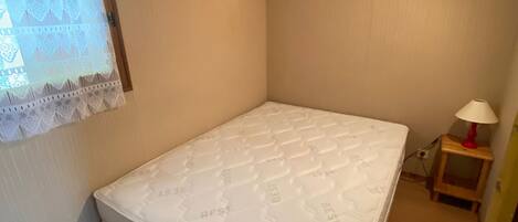 A small, single-bed room with a wooden ceiling, a bedside table with a lamp, and a window with lace curtains.
