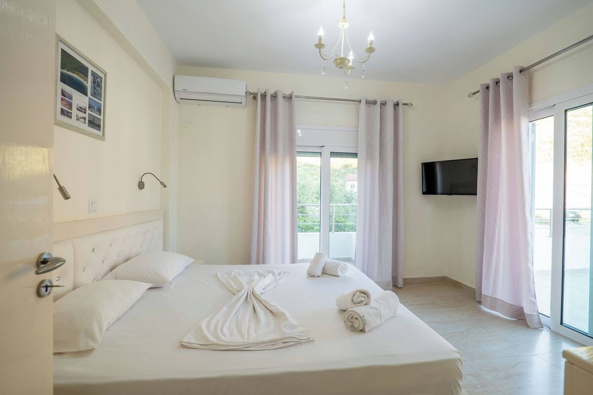 A hotel room with a bed, a chandelier, a flat-screen TV, and a view of greenery outside.