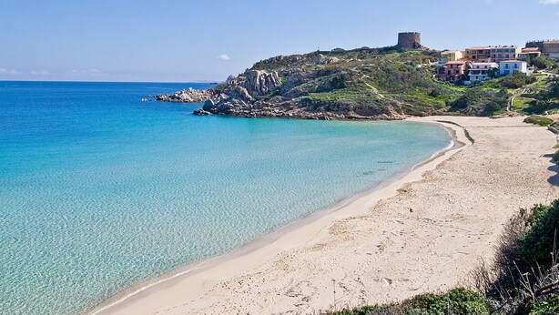Ein Küstenstrand mit klarem türkisblauem Wasser, einem Sandstrand und einem Hügel mit Gebäuden und einer Festung darauf.