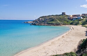 A coastal beach with clear turquoise waters, a sandy shore, and a hill with buildings and a fort on top.