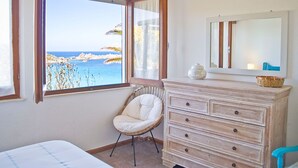 Ein Schlafzimmer mit großem Fenster und Blick auf den Strand, ein Korbstuhl, eine Holzkommode und ein Spiegel.