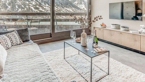 Sala moderna com uma grande janela com vista para montanhas, um sofá, uma mesa de centro com objetos decorativos e uma área de cozinha com uma TV.