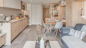Uma cozinha moderna com armários de madeira, uma sala de jantar com mesa posta para dois e um espaço de estar acolhedor com sofá e almofadas.
