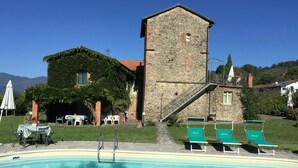 A pool area with lounge chairs, a stone building with a staircase, and a view of mountains. - La Casatorre di Ponticello. Casale di campagna con ampio giardino e piscina (Cantiere-ponticello)