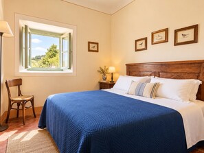 Une chambre avec un grand lit, une tête de lit en bois, une fenêtre donnant sur un paysage verdoyant, une chaise et des tableaux encadrés accrochés au mur.