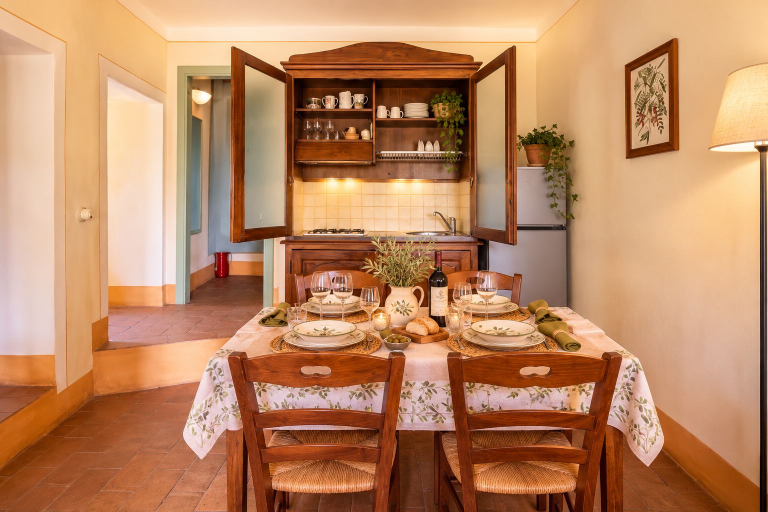 Zona pranzo con tavolo imbandito per quattro, sedie di legno, cucina sullo sfondo e una pianta in vaso appesa al muro.