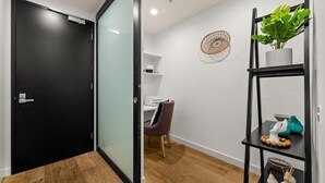 A modern interior with a black door, a glass partition, a wooden floor, a desk with a chair, a wall-mounted shelf with a plant, and a decorative wall piece. - StayCentral - Hawthorn East Elegance with Study (Hawthorn East)