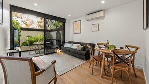 A modern living room with a sofa, dining table, and chairs. There is a large window with a view of greenery outside. - StayCentral - Hawthorn East Elegance with Study (Hawthorn East)