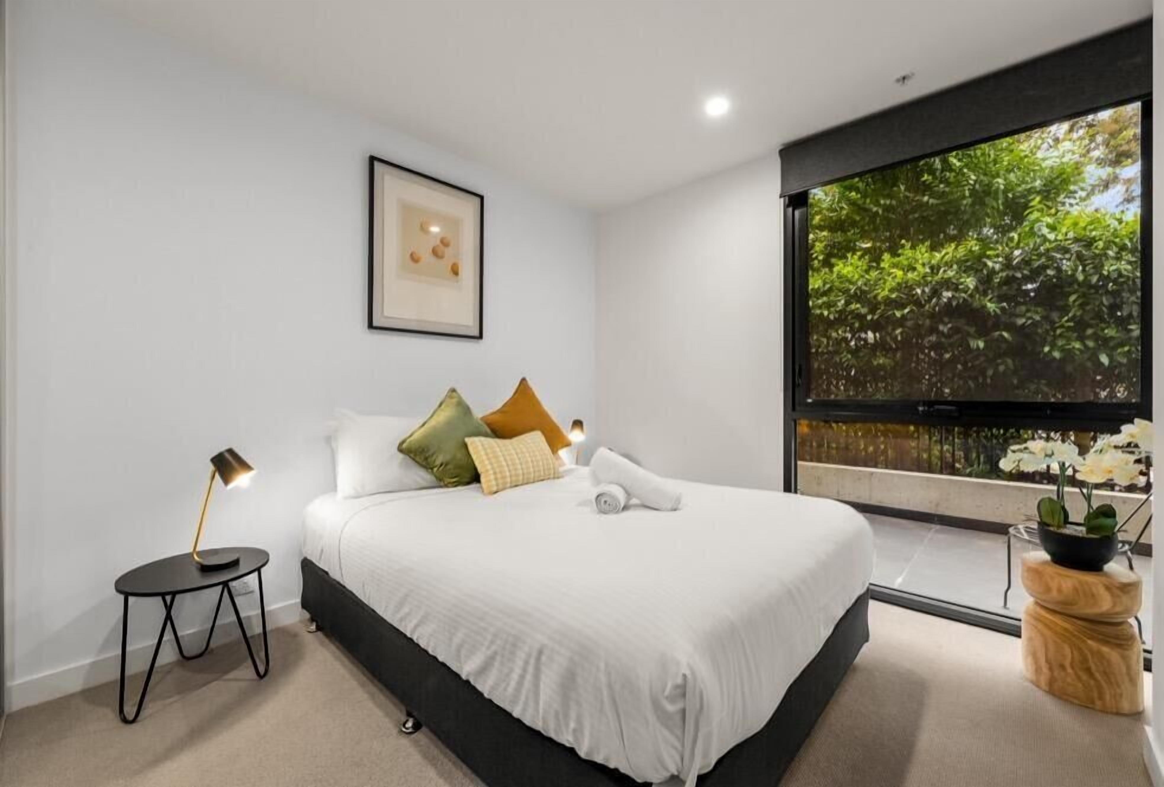 A modern bedroom with a large bed, a side table, a framed picture, and a window with a view of greenery.