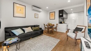 A modern living room with a black sofa, a coffee table, and a dining area with a table and chairs. There is a kitchen area with cabinets and a stove. - StayCentral - Hawthorn East Elegance with Study (Hawthorn East)