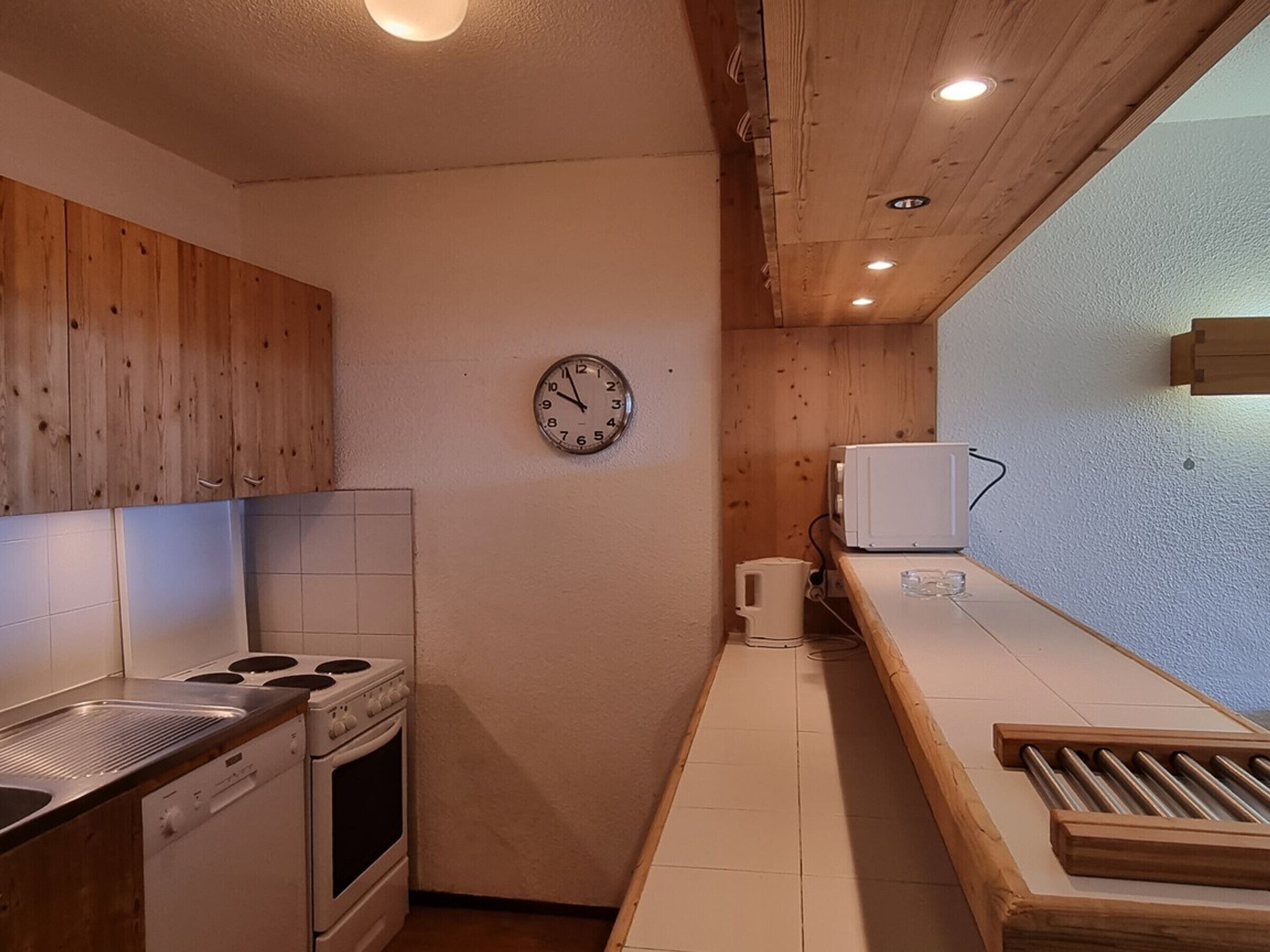 Une cuisine de taille réduite, avec des meubles de rangement en bois, une cuisinière, un four à micro-ondes et une horloge au mur.