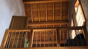 Una habitación con techos altos, vigas de madera, un balcón con barandilla y vistas al exterior.
