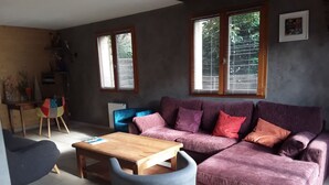 Una sala de estar con un sofá morado, una mesa de centro de madera y un sillón azul. Hay dos ventanas con persianas, un radiador y una pequeña mesa con una planta.