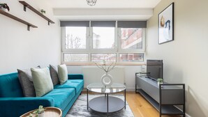 A modern living room with a teal sofa, a glass coffee table, a wooden side table, and a grey rug. - Amazing Flat in Battersea (London)