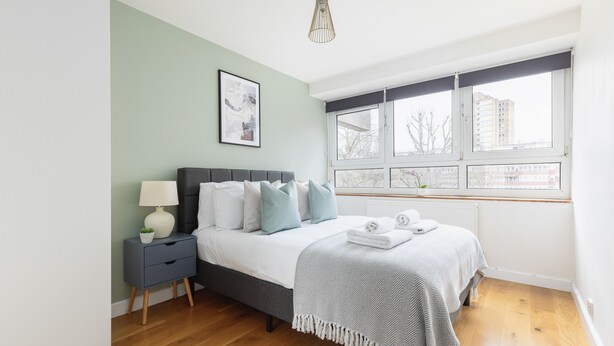 A bedroom with a bed, bedside table, lamp, and a window with a view of buildings. - Amazing Flat in Battersea (London)