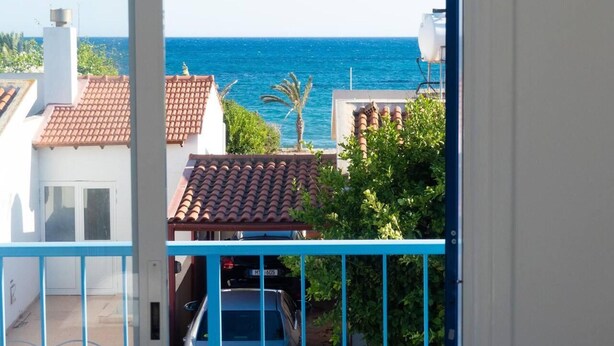 Vista de varanda com vista para a costa, mar azul cristalino, uma palmeira e edifícios com telhados de terracota.