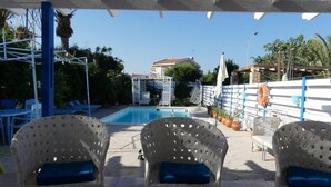 Uma área à beira da piscina com três cadeiras de vime com almofadas azuis, um vaso branco decorativo e vista clara para a piscina.