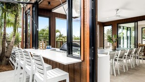 Un área de comedor al aire libre con sillas blancas y una mesa larga, rodeada de paredes de madera y ventanas grandes.