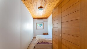 A narrow hallway leading to a bedroom with a wooden ceiling, a bed with a white and purple bedspread, and a small bedside table.