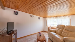 A living room with a wooden ceiling, a sofa, a TV on a wooden stand, a small table, and a window with curtains.