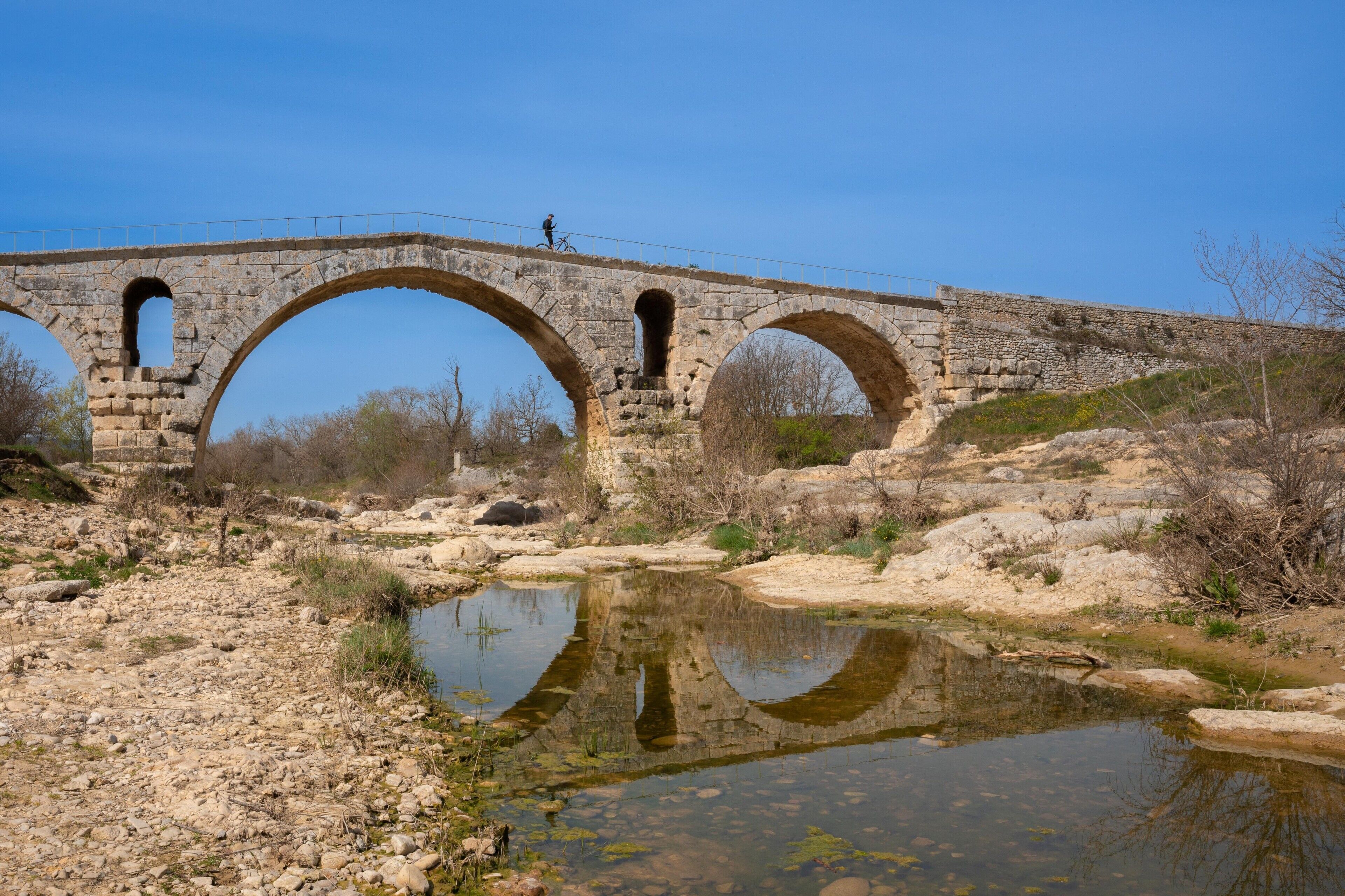 A stone bridge with three arches over a body of water, surrounded by rocky terrain and sparse vegetation.
