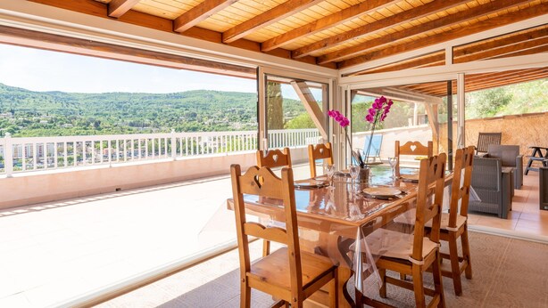 A wooden dining table with chairs, a glass table, and a view of green hills.
