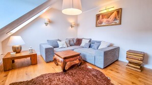 A cozy loft bedroom with a large L-shaped gray sofa, a wooden coffee table, a fluffy brown rug, and a small wooden side table with a lamp. - Luxurious apartment * Air conditioning * Parking lot (Coburg)