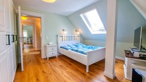 A bedroom with a sloped ceiling, a skylight, a bed with blue bedding, a bedside table, and a TV stand with a monitor. - Luxurious apartment * Air conditioning * Parking lot (Coburg)