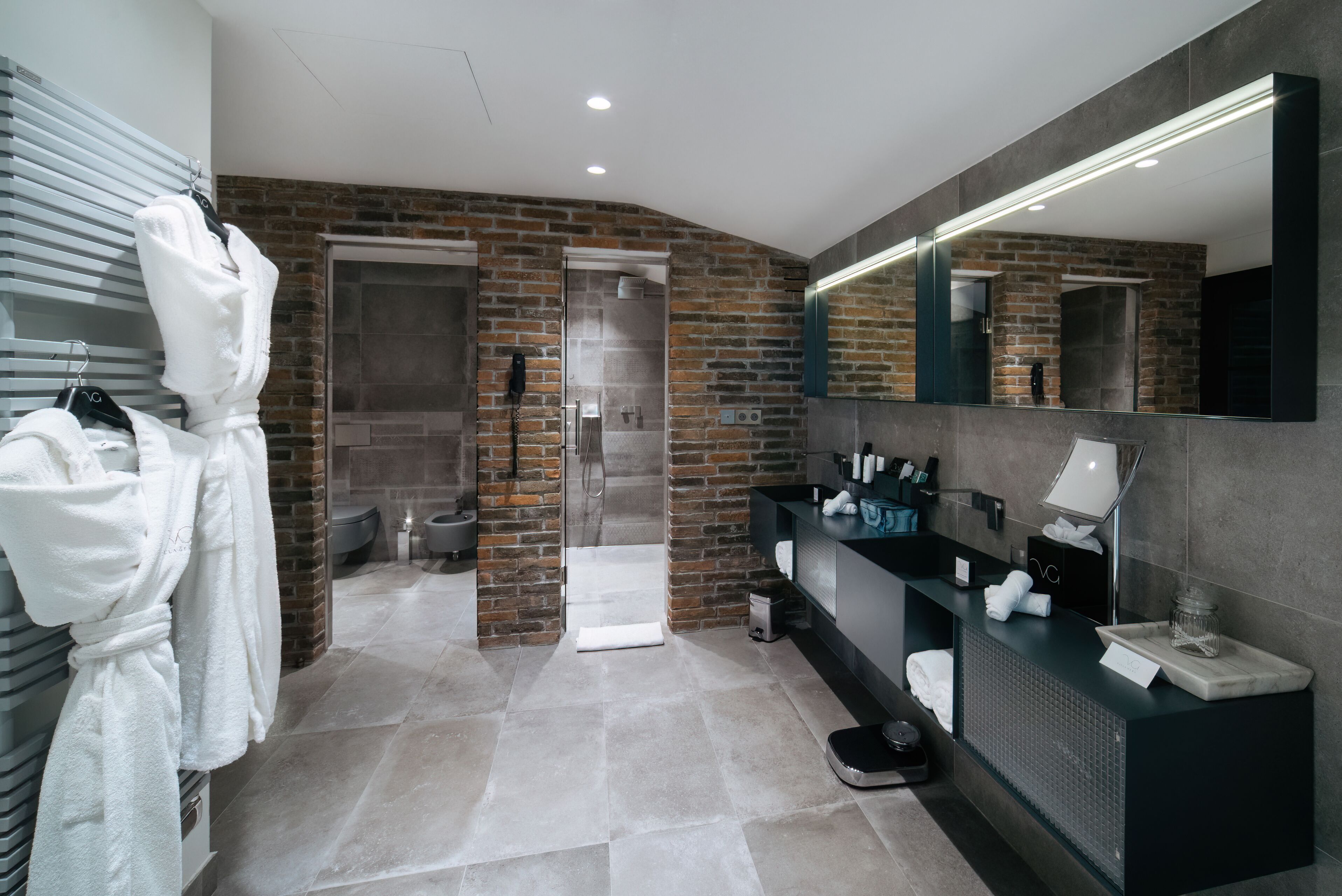 Une salle de bain moderne avec un mur en briques, un grand miroir, un meuble-lavabo avec des articles de toilette et un espace douche.