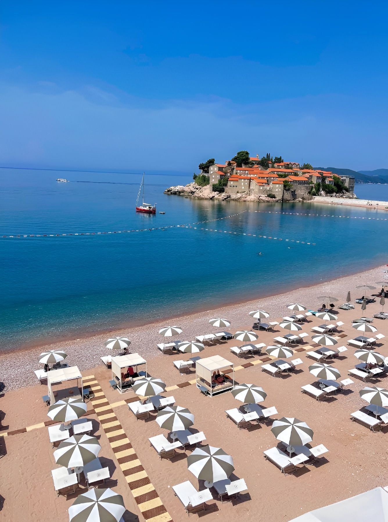 Ein Strand mit vielen Sonnenschirmen und Liegestühlen, einem klaren blauen Meer und einem historischen Gebäude auf einem Felsen.