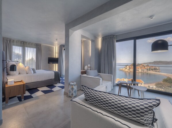 A modern hotel room with a large bed, a sofa, a small table, and a view of the sea.