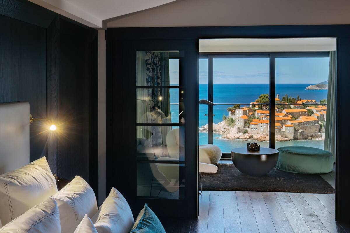 A modern bedroom with a large bed, a seating area with a round table, and a balcony with a view of the sea and buildings.